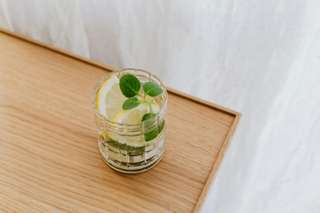 A clear glass of water with lemon slices on a wooden table surrounded by fresh fruits